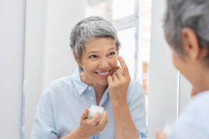 A cheerful mature woman applying face cream while looking in the mirror, enjoying her skincare routine.