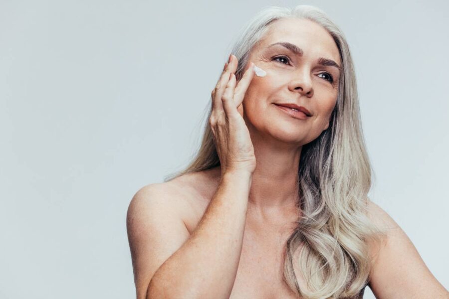 Mature woman with silver hair applying facial cream, looking serene against a light background.